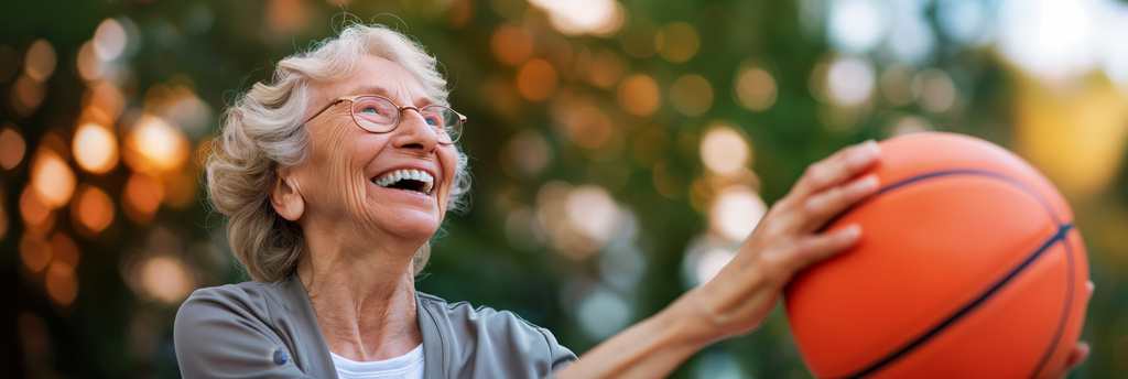 An elderly person enjoying a game of basketball outdoors with an orange ball, shown during a warm, golden sunset, emphasizing vitality and joy.
